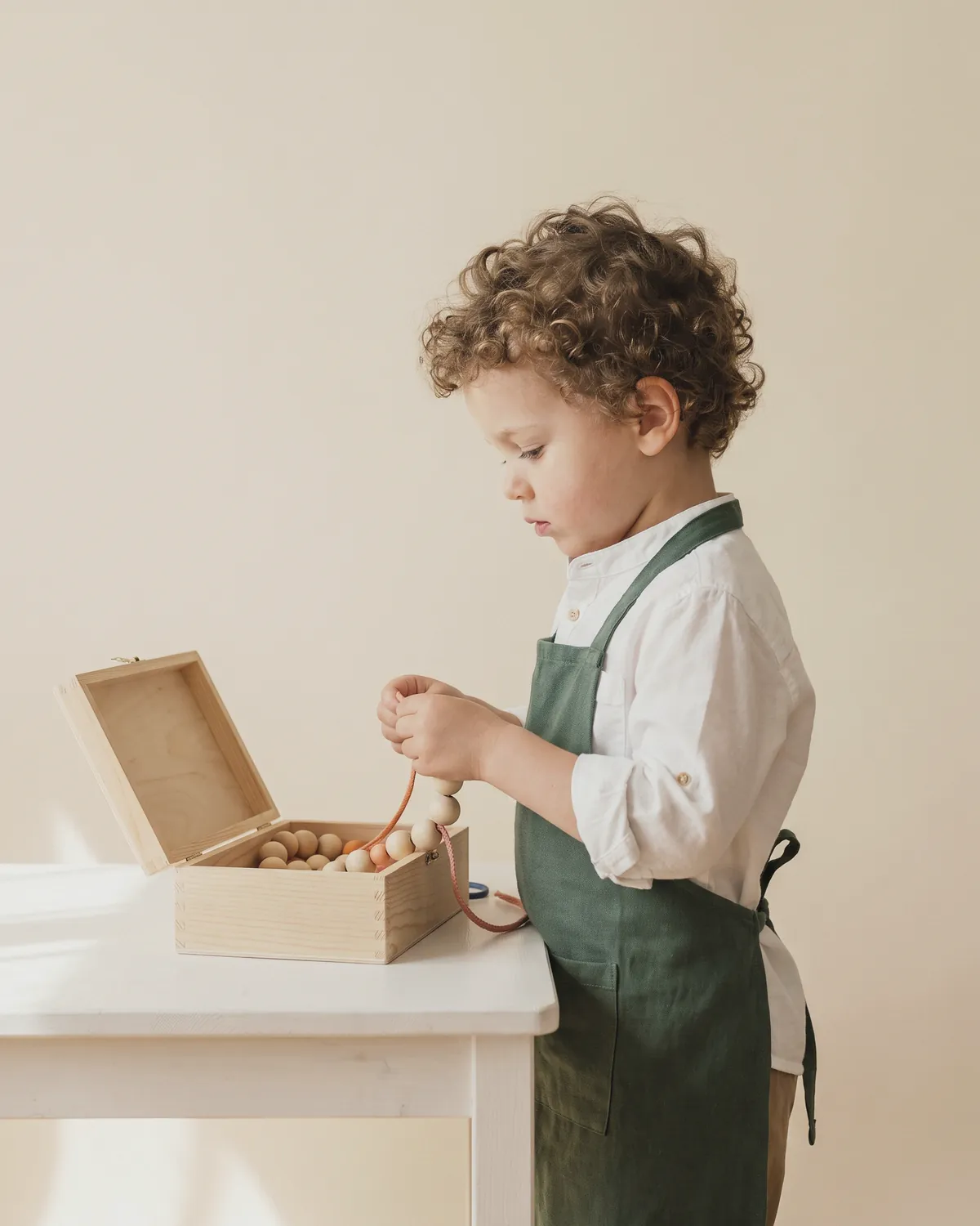 Illustration: a curly-haired boy in a white shirt and green apron threading wooden beads from an open wooden box on a white table