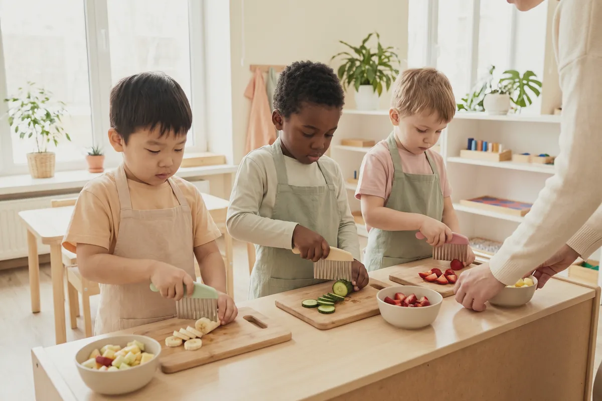 Illustration: three preschoolers in aprons cutting banana, cucumber and strawberries with wavy child-safe cutters on wooden boards at a low classroom counter