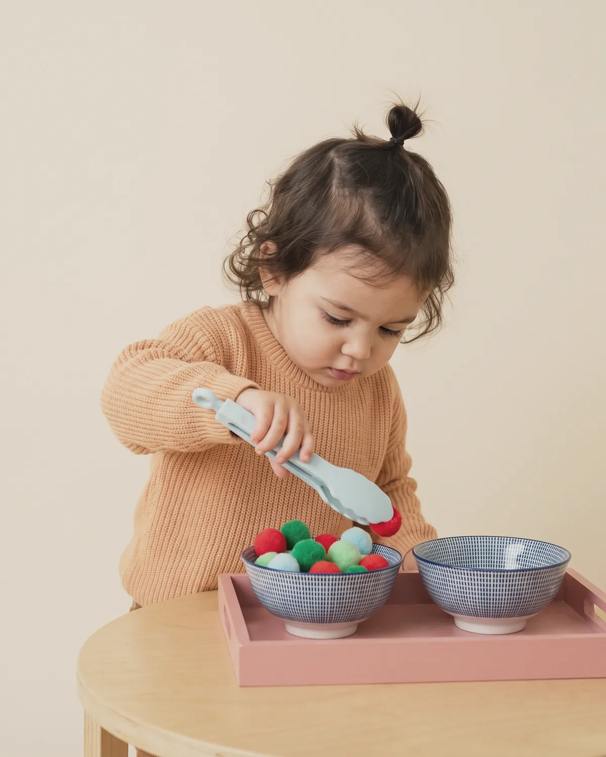 Illustration: a toddler in an apricot sweater using light-blue tongs to lift a pom-pom from a patterned bowl on a pink tray