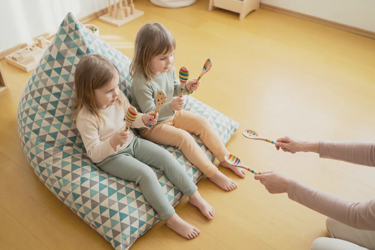 Illustration: two little girls on a triangle-patterned bean bag holding painted wooden spoons while an adult's hands hold out two more