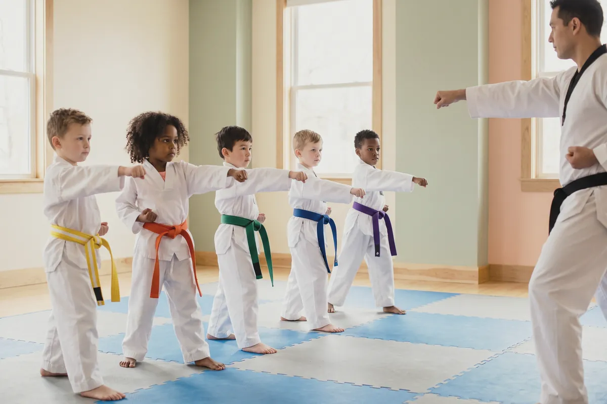 Illustration: five young children in white taekwondo uniforms with coloured belts throwing a front punch in a line while a black-belt instructor demonstrates in a bright mat room