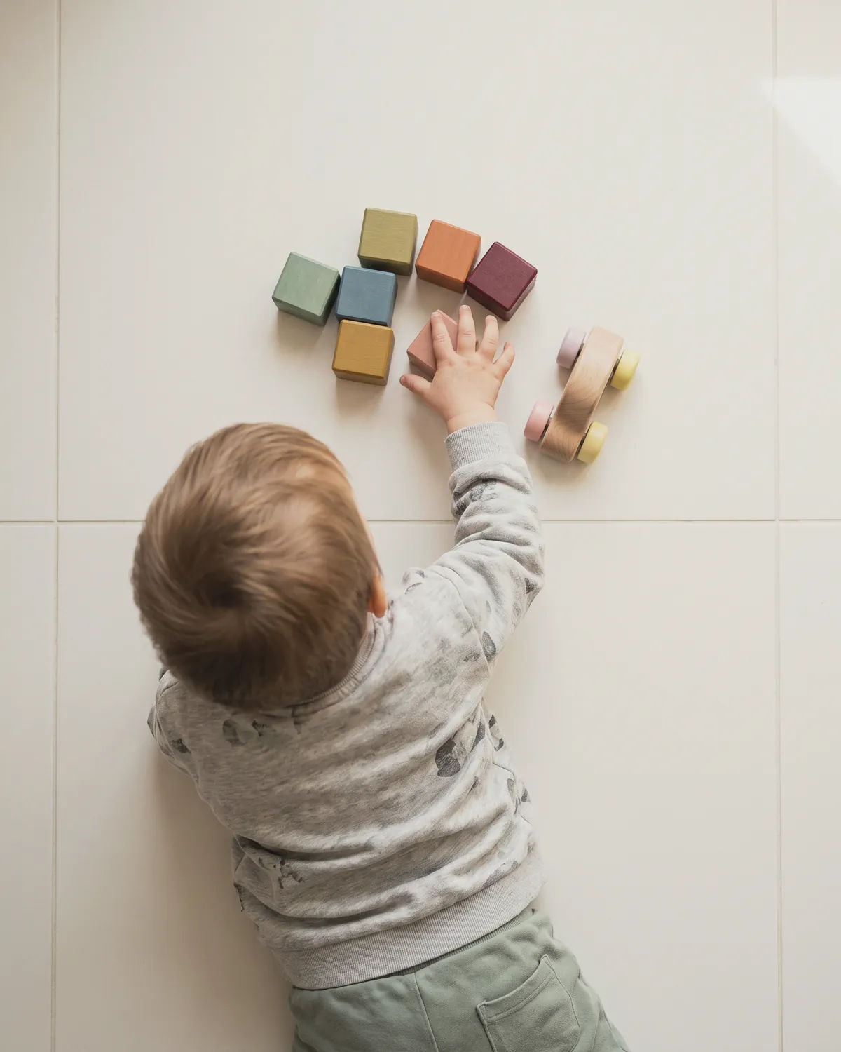 Illustration: a toddler in a grey sweatshirt lying on a white tile floor, reaching toward coloured wooden blocks and a wooden toy car, seen from above
