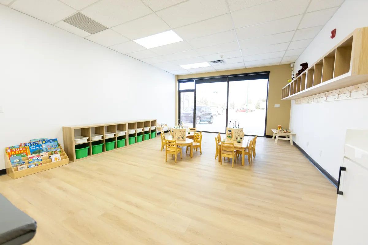 A bright, empty classroom with a rack of picture books, low cubbies of green bins, small wooden tables and a wall of windows