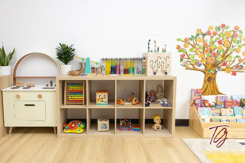 A low shelf of sensory bottles, dolls and toys beside an autumn tree wall decal and a play kitchen