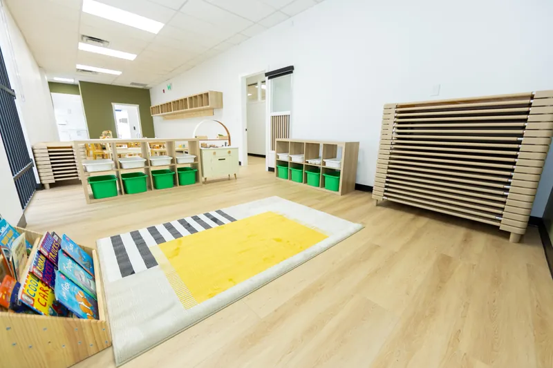 A yellow and grey rug on a wooden floor beside a bin of picture books, with cubby shelves of green bins and a stack of nap cots