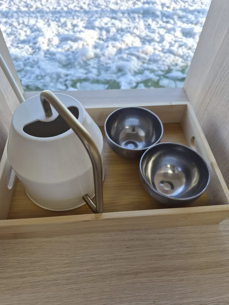 A wooden tray holding a white watering can and two steel bowls on a table by a window