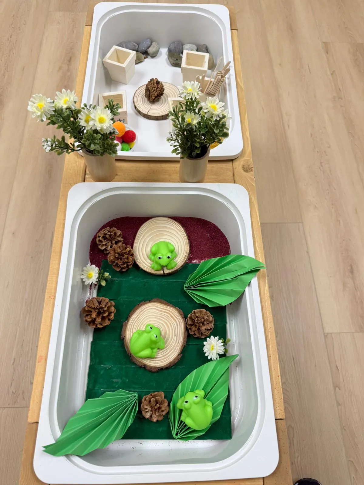 A tub filled with green felt, wooden slices, pinecones and toy frogs, next to a tub with wooden cups, stones and daisies