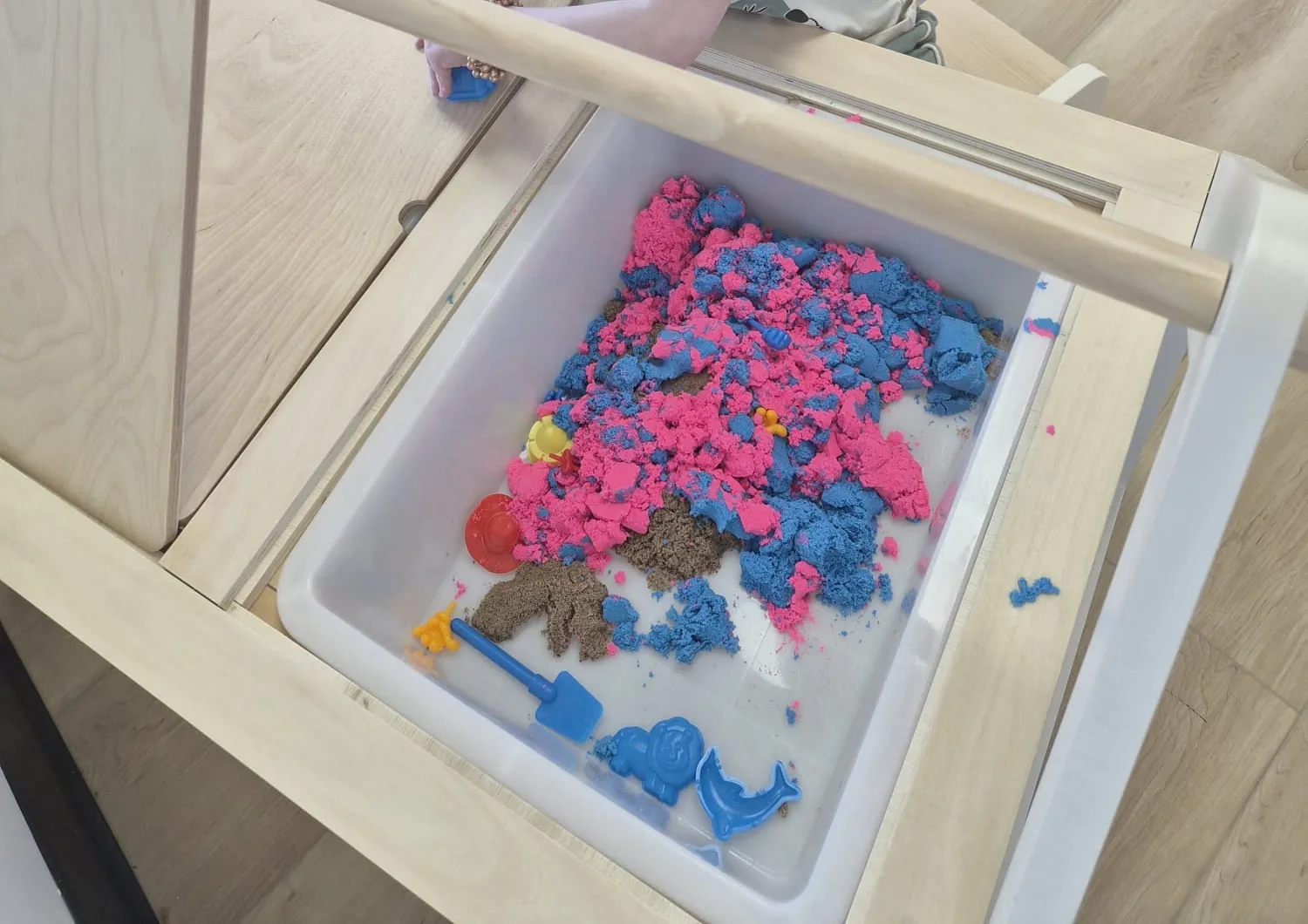 A bin of pink and blue kinetic sand with a small shovel and dolphin moulds on a wooden sensory table