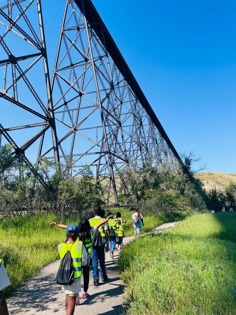 Children in yellow safety vests walking single file, seen from behind, along a path beneath a tall steel railway bridge