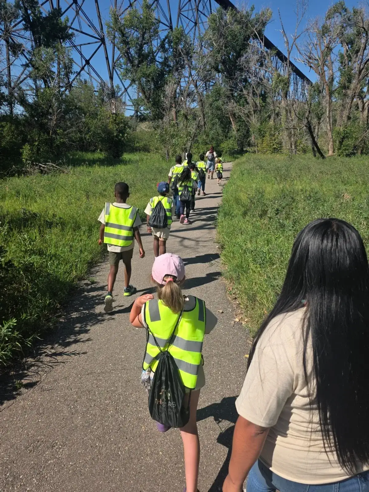 Children in yellow safety vests with drawstring backpacks walking along a paved trail lined with tall grass, seen from behind