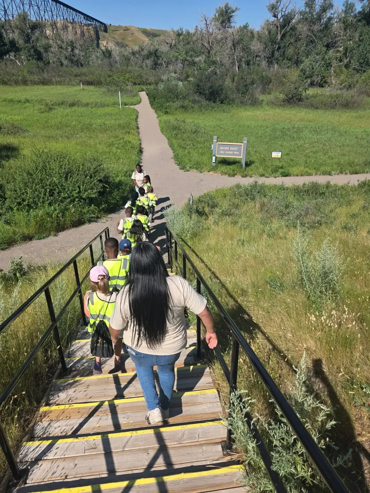 A line of children in yellow safety vests walking down a wooden staircase toward a grassy trail, seen from above and behind