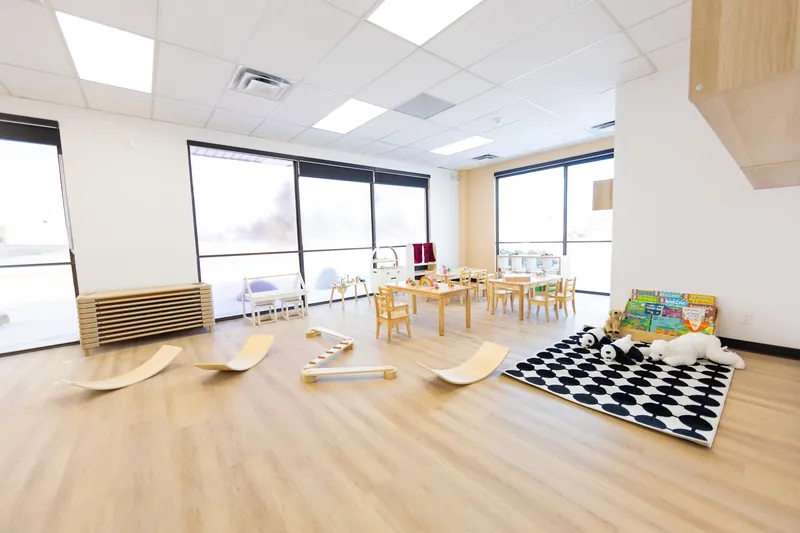 A bright, empty classroom with wooden balance boards on the floor, a black and white rug, small tables and chairs, and a wall of windows