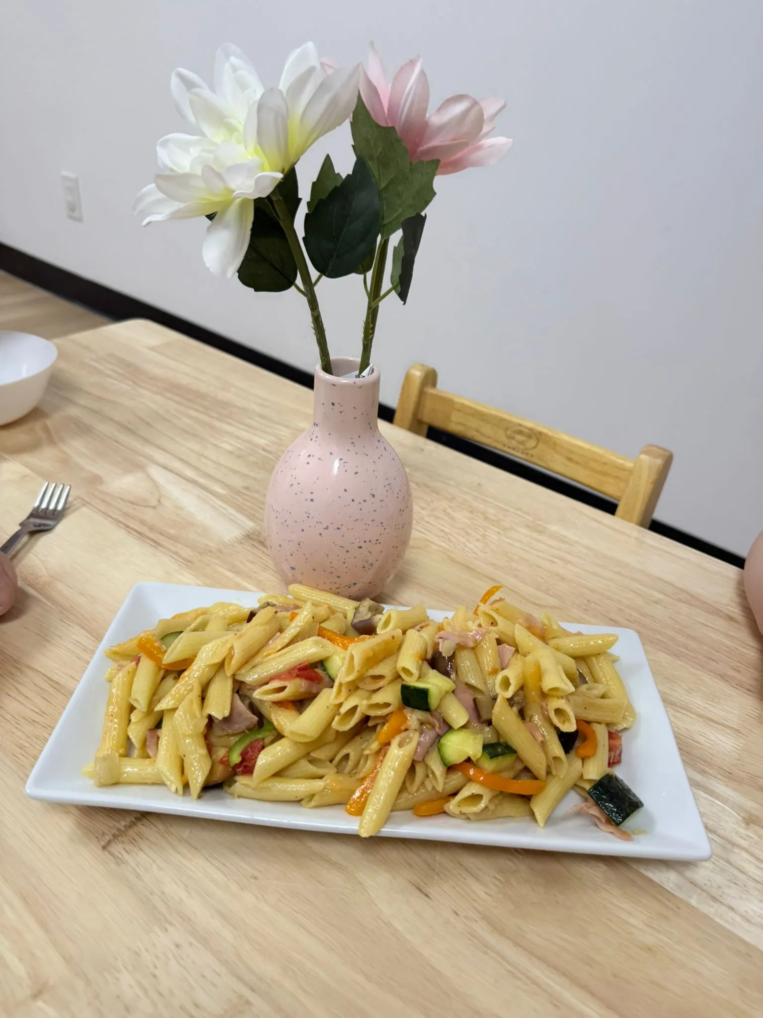 A white rectangular plate of penne pasta with sliced vegetables beside a small vase of pink and white flowers on a wooden table