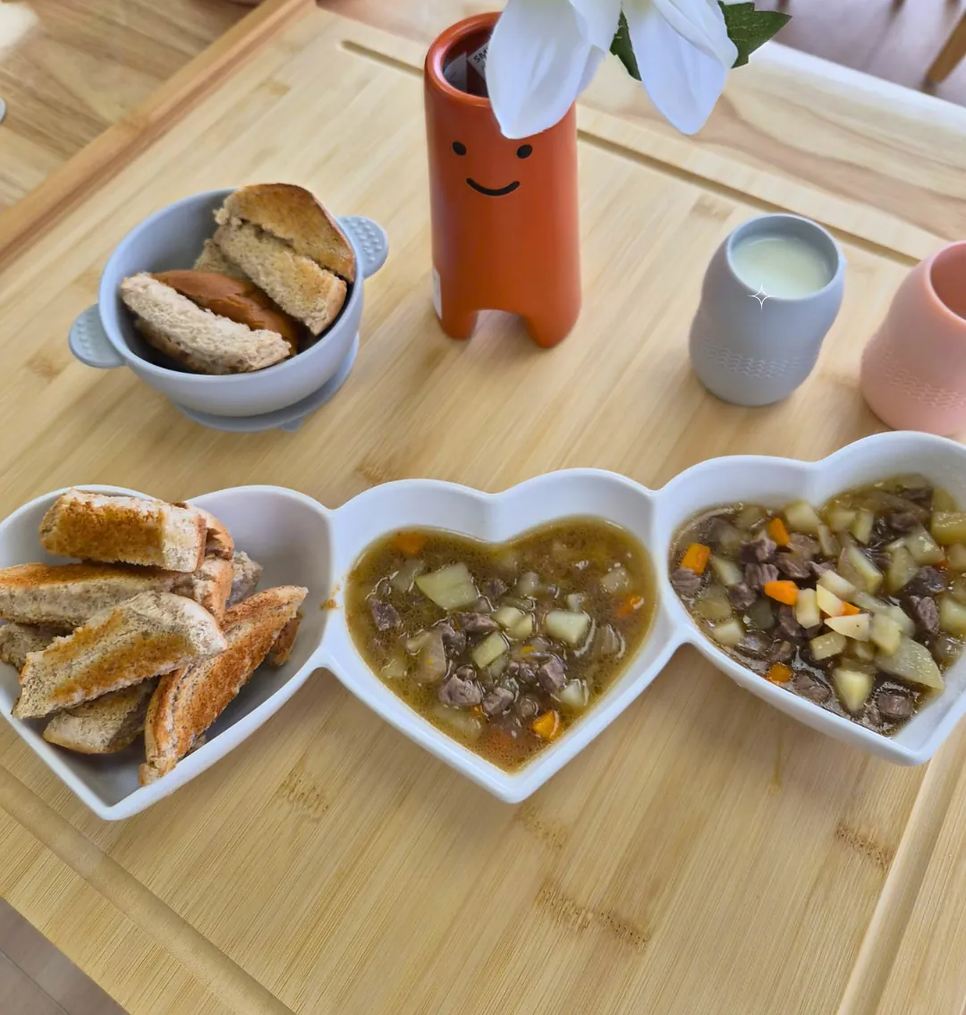 A heart-shaped three-part dish of beef and vegetable stew and toast, with a bowl of toast, a cup of milk and a vase of flowers on a wooden table