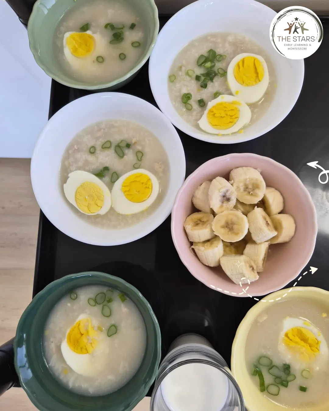 Bowls of rice porridge topped with halved boiled eggs and green onion, a bowl of sliced banana and a glass of milk, seen from above