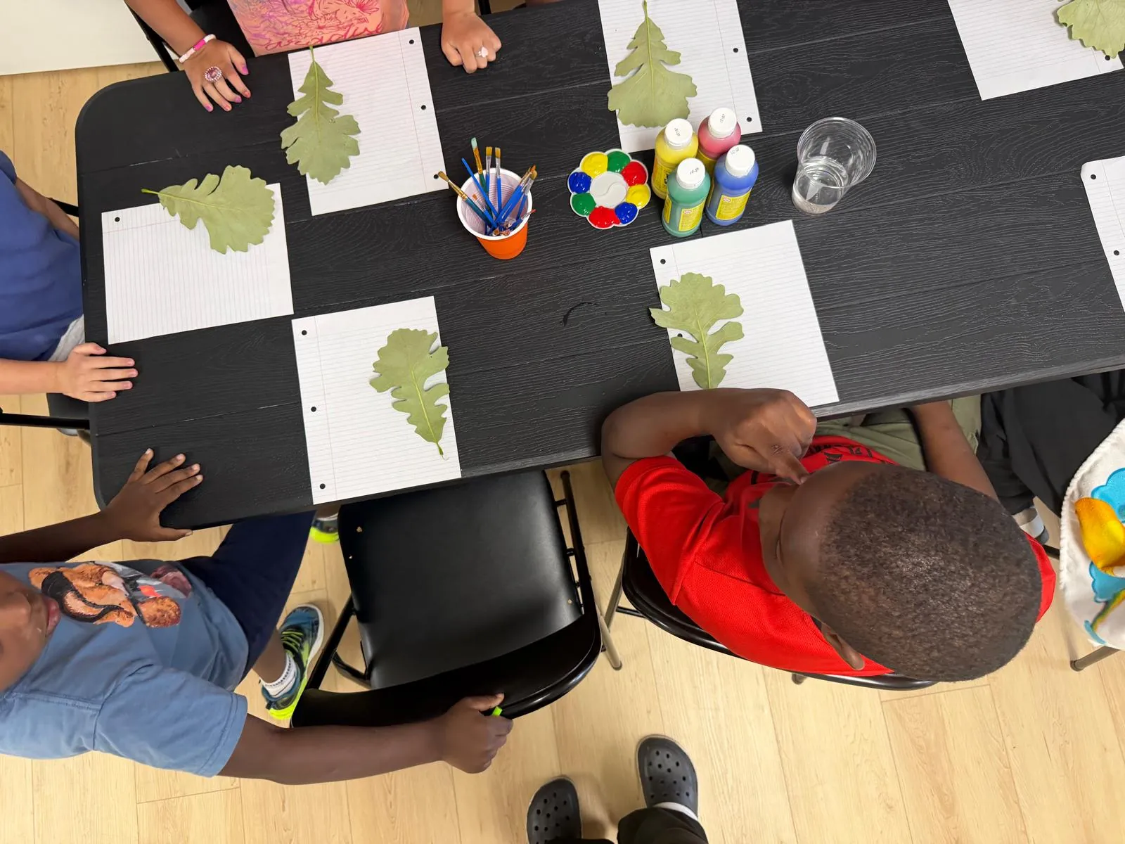 A table seen from above with sheets of paper, oak leaves, paint bottles and brushes, with children's hands around it