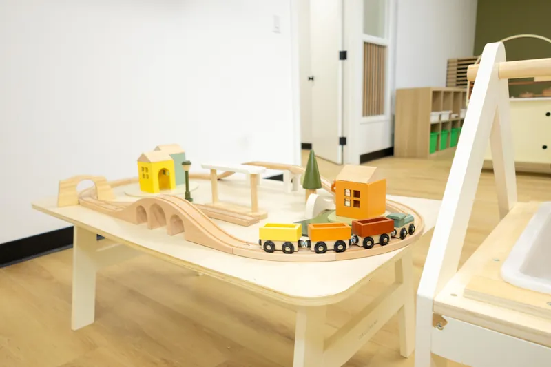 A wooden toy train set with a bridge, small painted houses and a tree on a low pale table